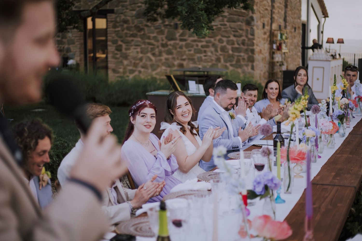 Wedding reception dinner with guests seated at a long table near stone walls.