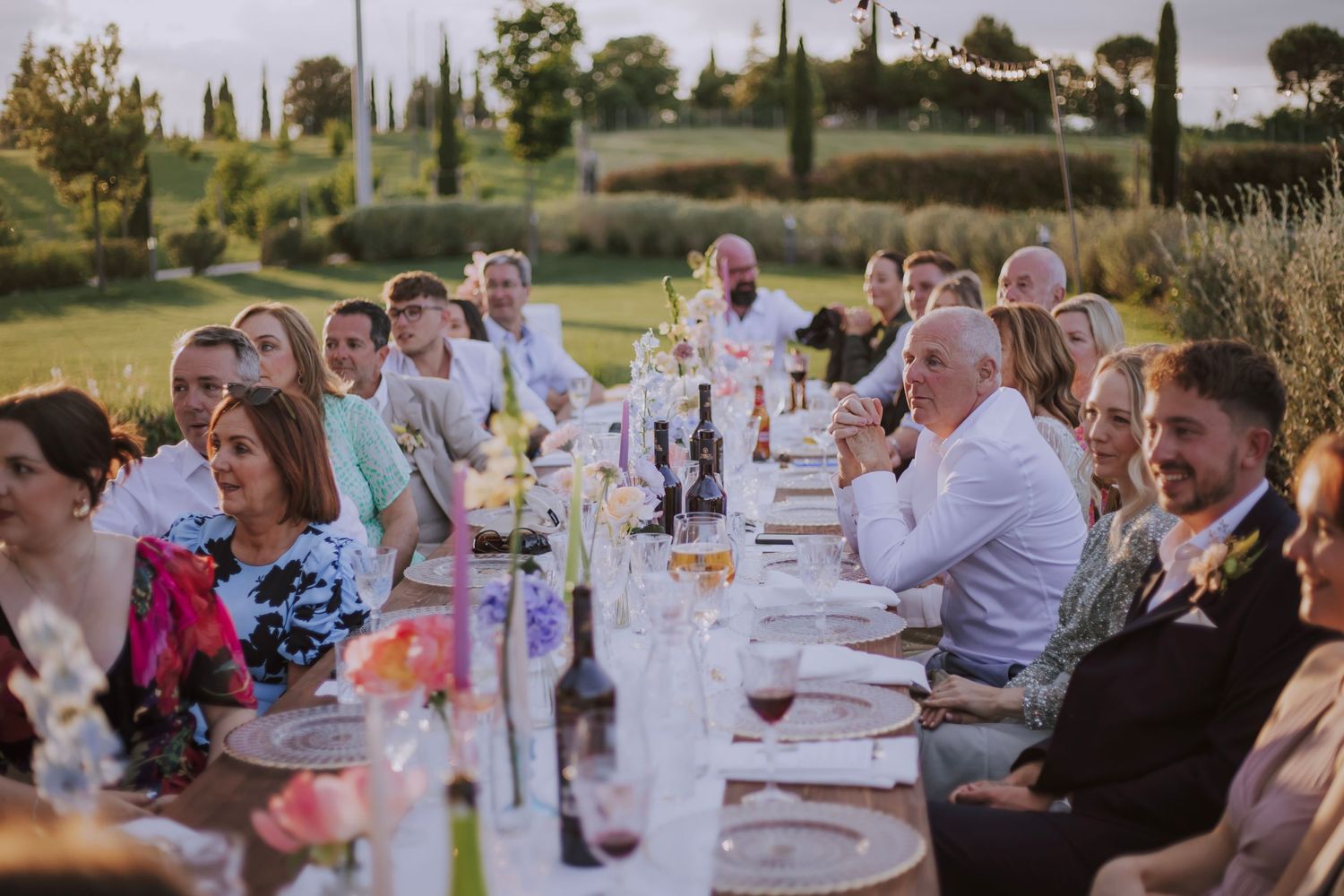 Outdoor wedding reception with guests dining at a long white-clothed table.