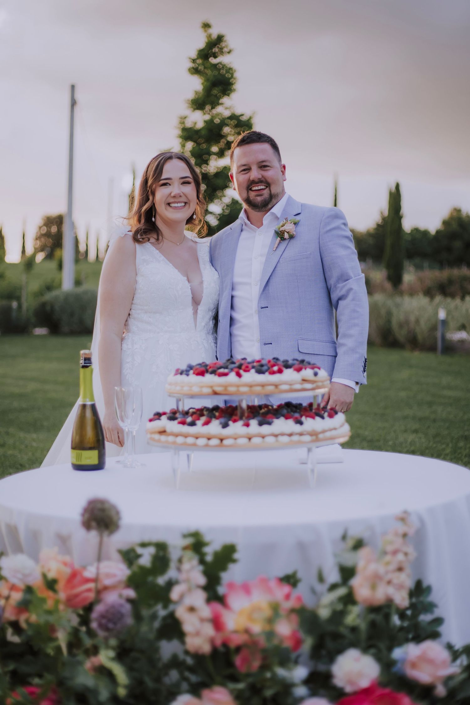 Wedding couple stands behind a tiered berry-topped cake at their outdoor reception with floral decorations.