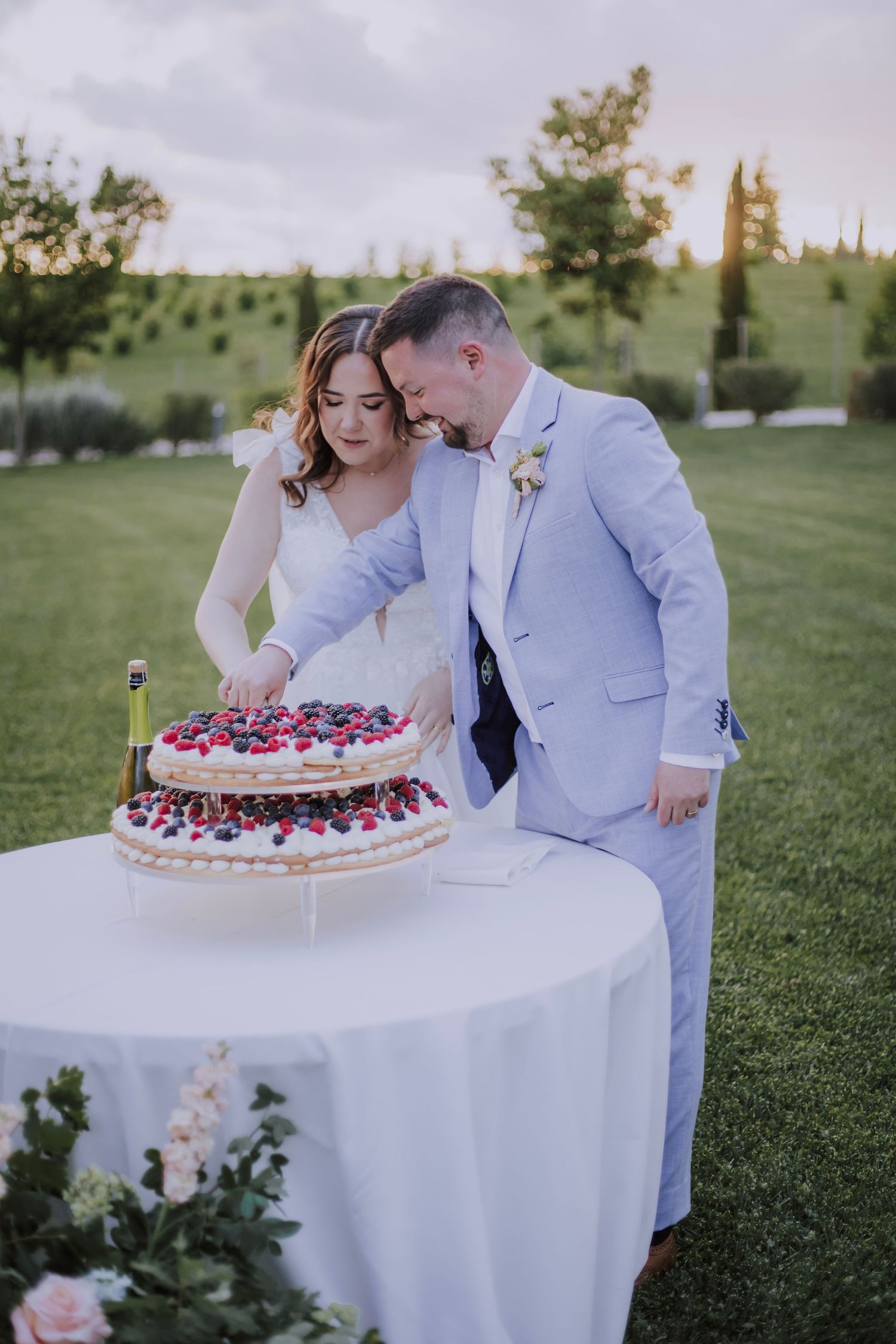 Newlyweds lean in together while cutting their two-tiered berry-decorated wedding cake outdoors.