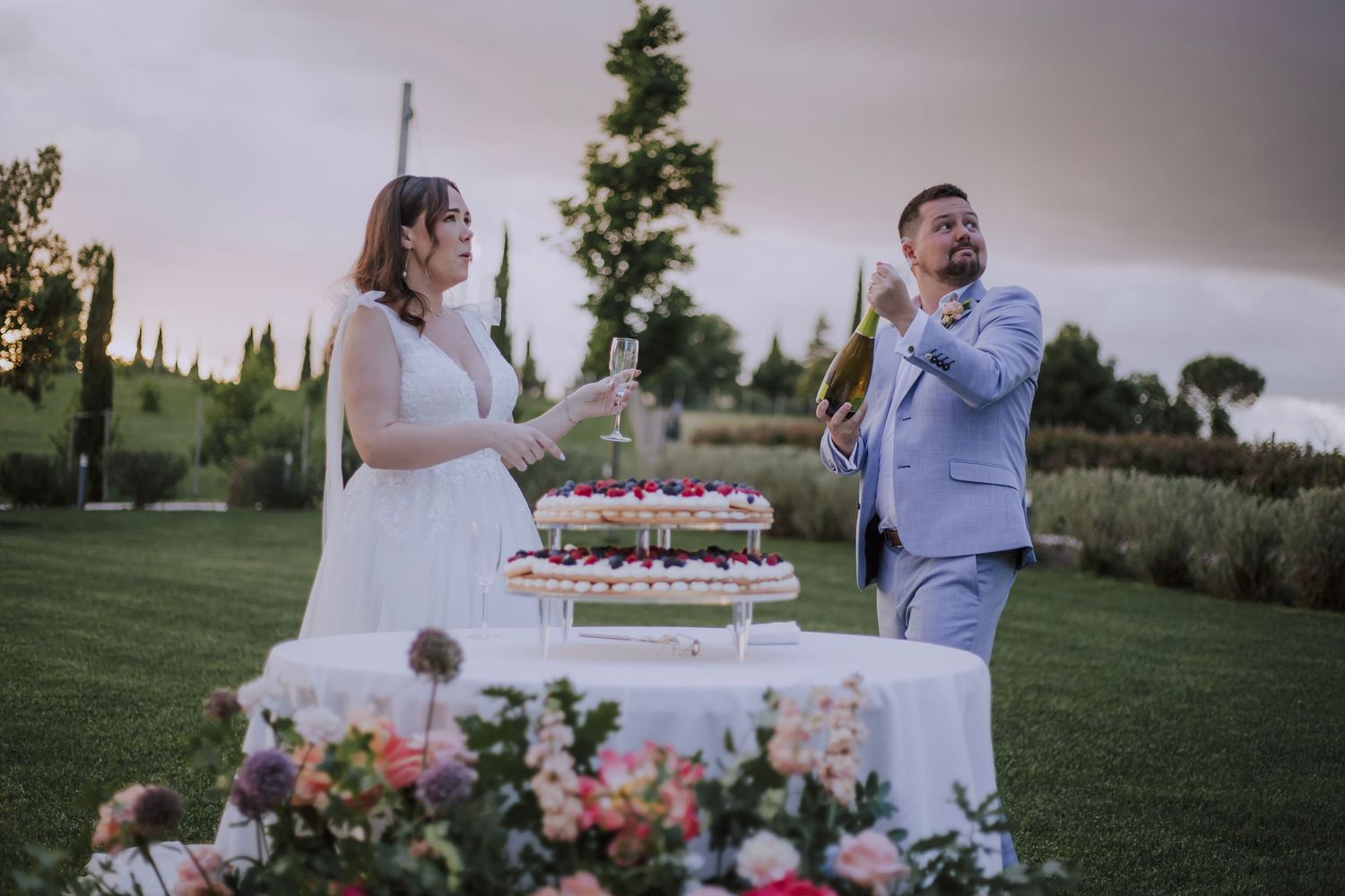 Wedding couple laughs while sharing cake at their outdoor reception surrounded by flowers.