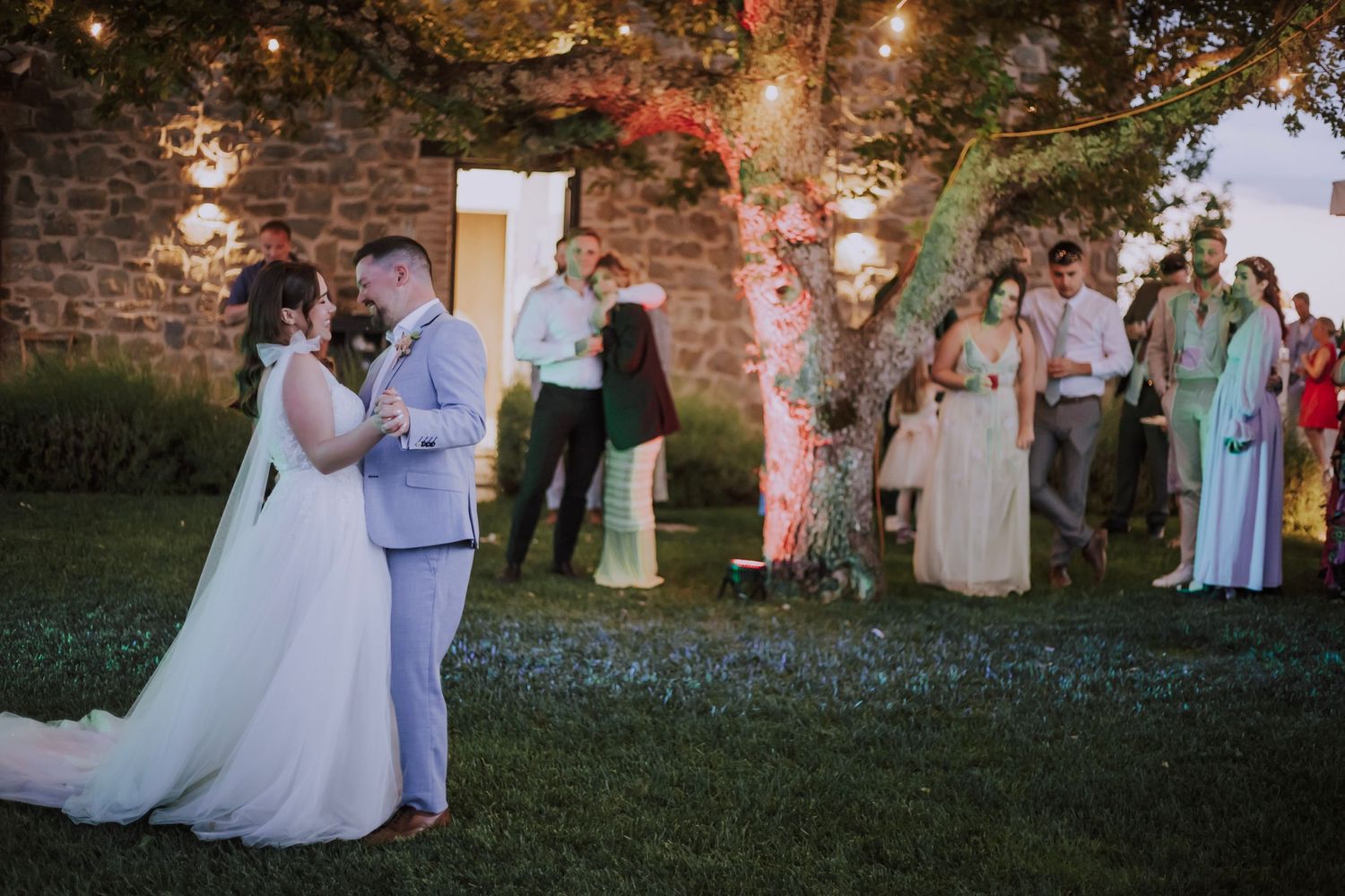 Wedding couple shares their first dance at dusk under string lights in a garden setting.