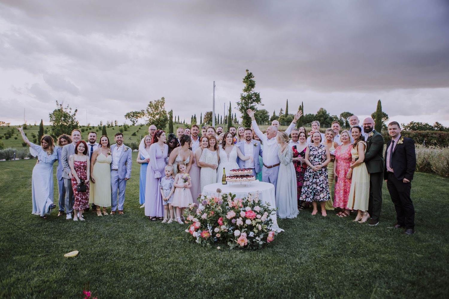 Large wedding party poses for a group photo on a lawn with floral arrangements.