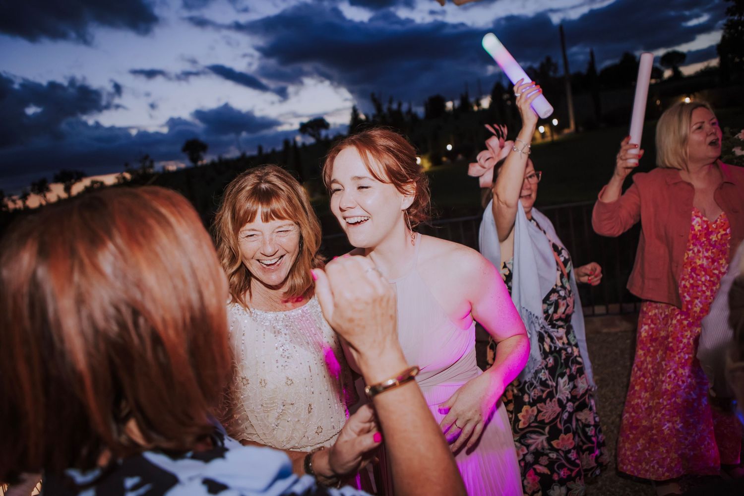 Wedding guests dance and celebrate at a twilight outdoor reception with colorful lighting.