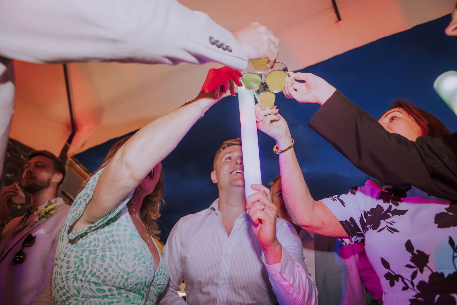 Wedding party toasting and raising glasses together during celebration under outdoor tent.
