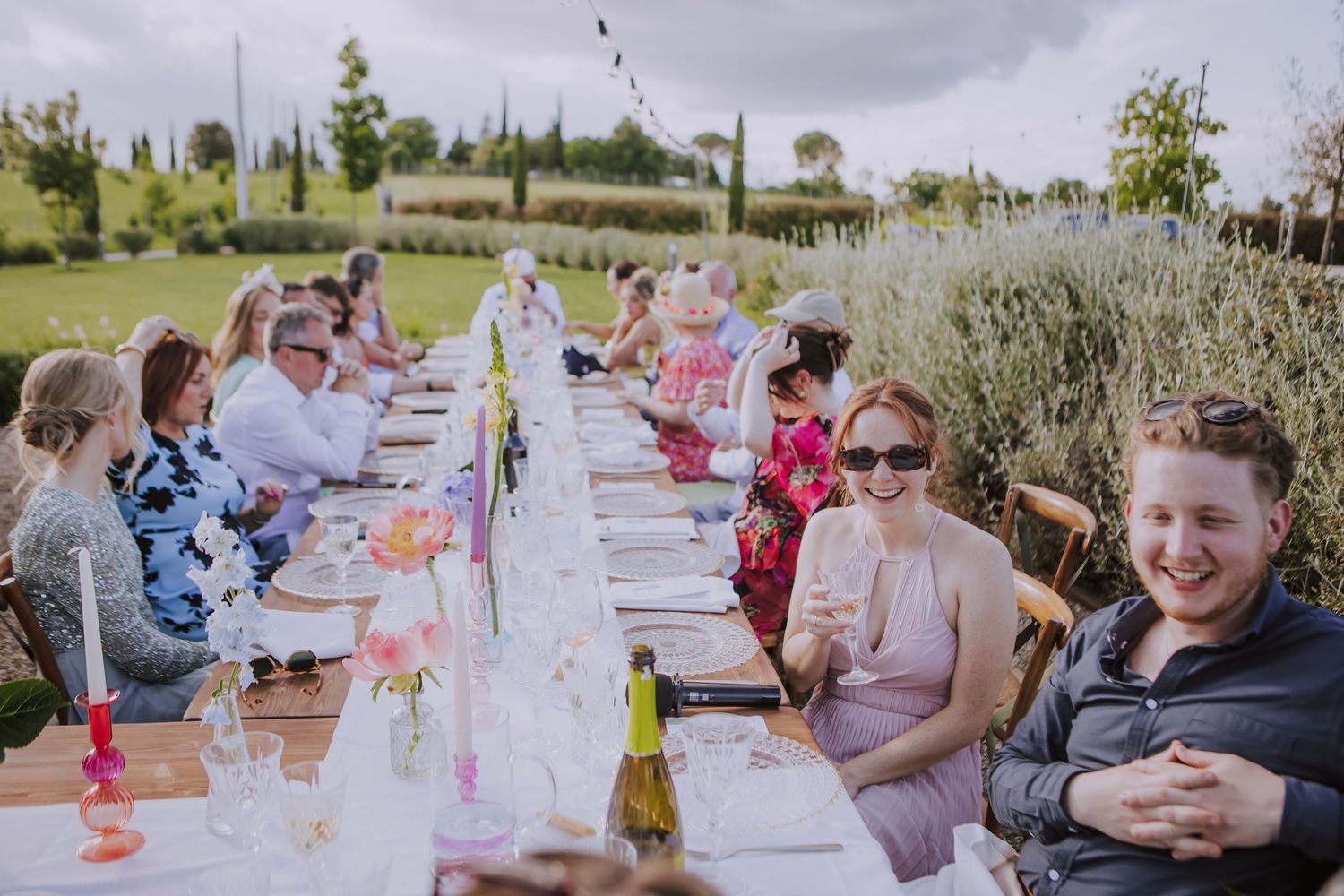 Wedding guests enjoying an outdoor dinner reception at a long table under cloudy skies.