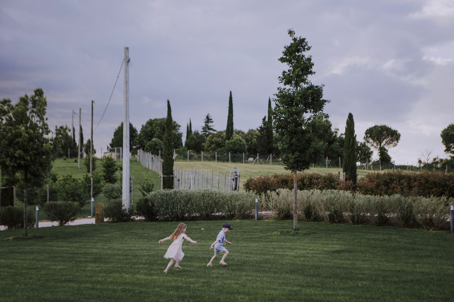 Children running and playing on a green lawn with cypress trees in the background.