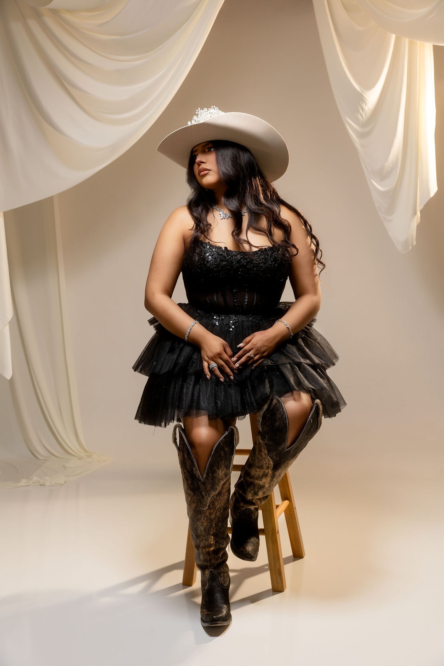 Woman in a black sparkly tulle mini dress, cowboy boots and cream cowboy hat sits on a wooden stool before draped ivory curtains.