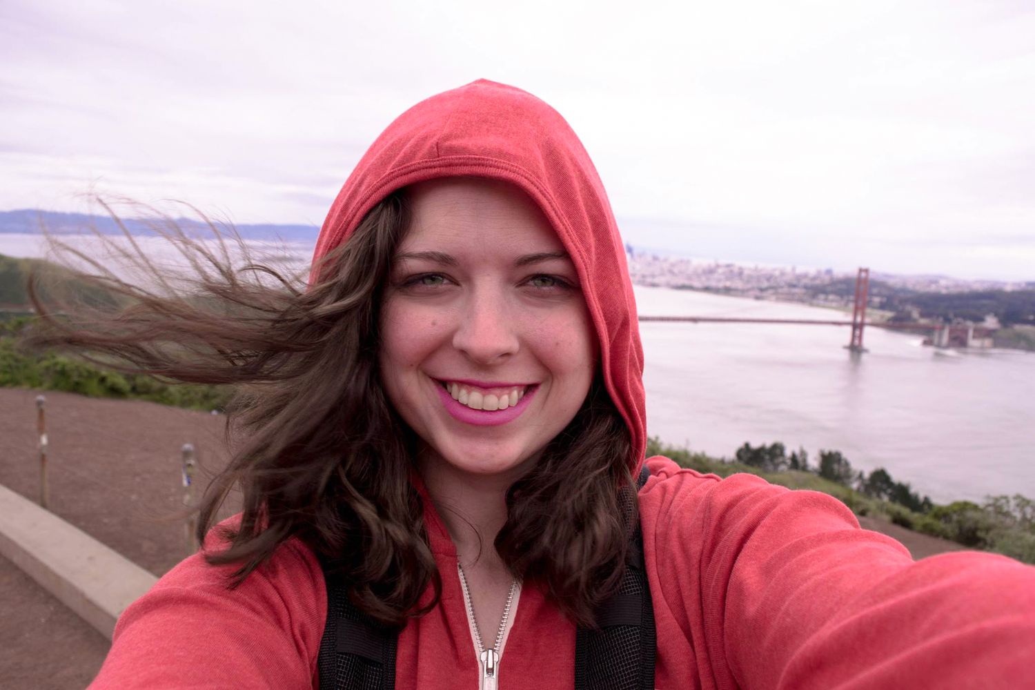 Smiling woman in red hoodie takes selfie with Golden Gate Bridge and San Francisco Bay in background.