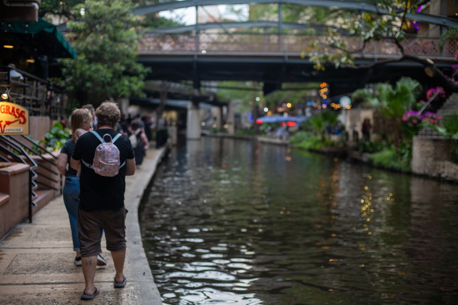 Two people walking along the scenic San Antonio River Walk canal path with a bridge and lush greenery in the background.