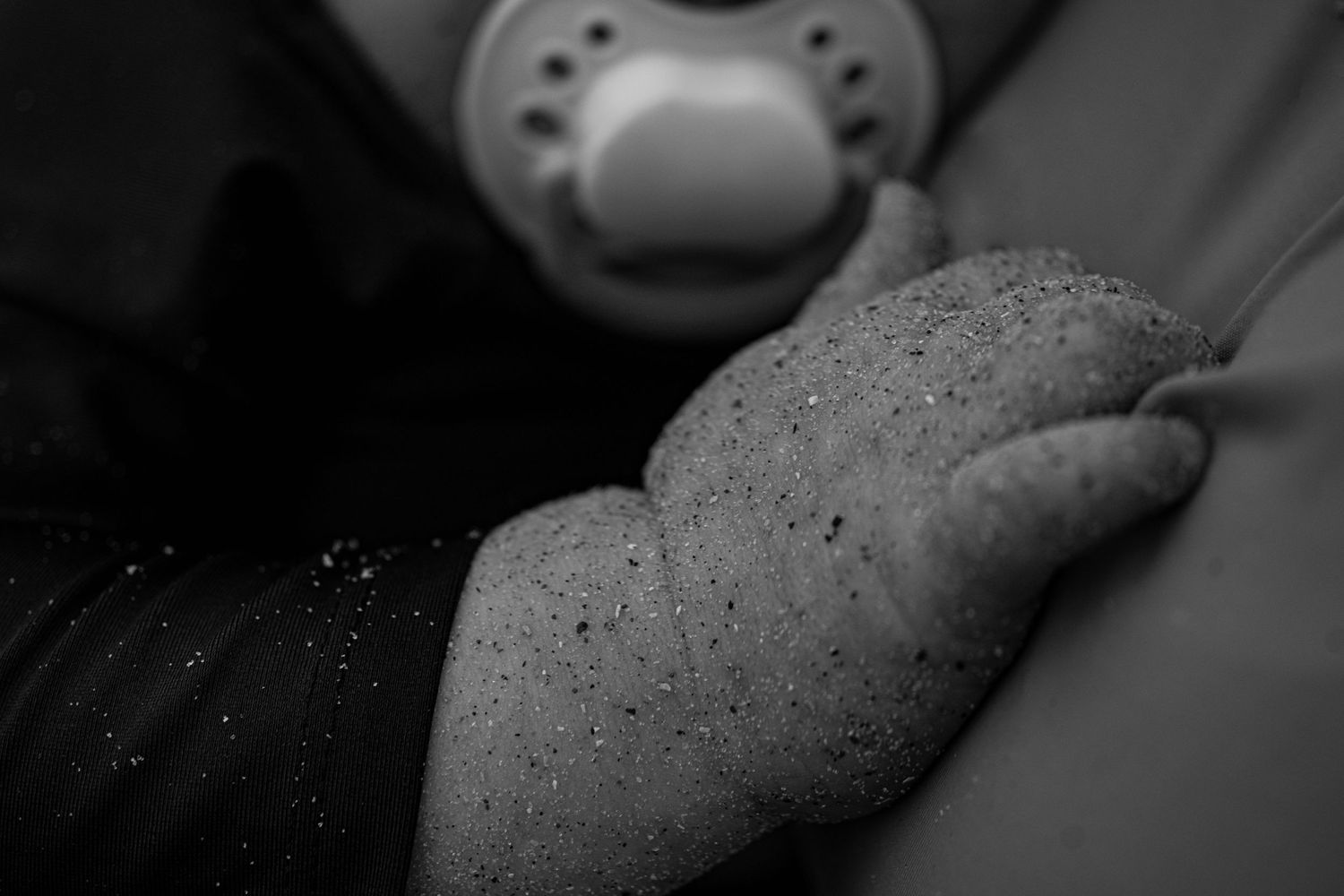 Close-up black and white photo of a sleeping baby's tiny hand with a pacifier in the background.