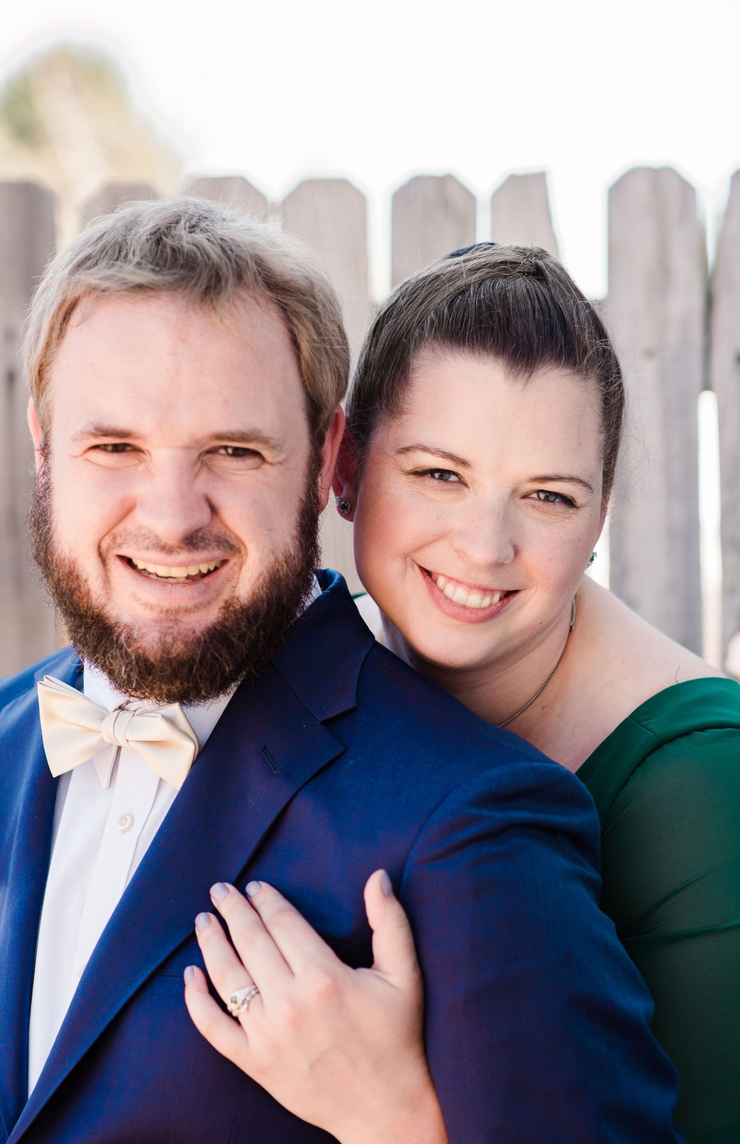A smiling couple poses outdoors near a wooden fence, man in blue suit with bow tie, woman in green dress wearing a ring.