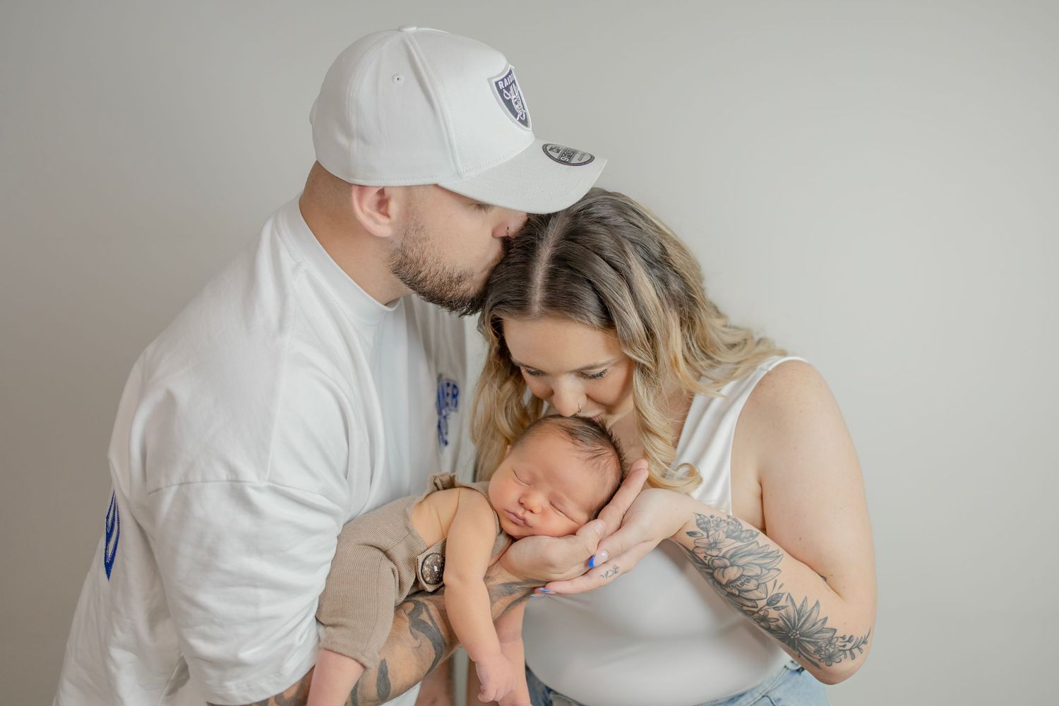 Parents in white outfits tenderly kiss their sleeping newborn baby during a studio portrait session.