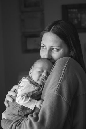 Black and white photo of a young mother tenderly holding her sleeping newborn baby close to her chest indoors.