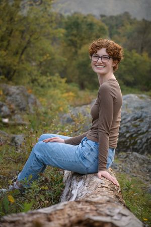 Young woman with curly red hair sitting on log in autumn nature setting.