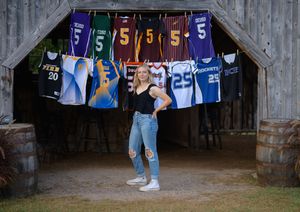 Student poses under sports jerseys hanging in barn doorway for senior photos.