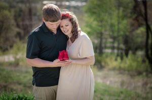 Couple sharing a tender moment outdoors, woman in beige dress with red flower crown holding a red rose bouquet.