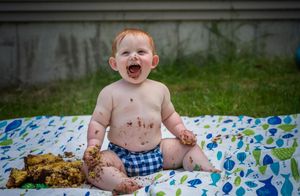 Messy baby laughing outdoors on blanket with chocolate cake smeared on face and hands.
