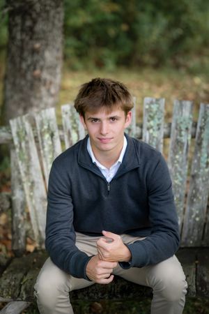 Young teen boy in gray quarter-zip sweater sitting on rustic wooden fence in autumn forest.