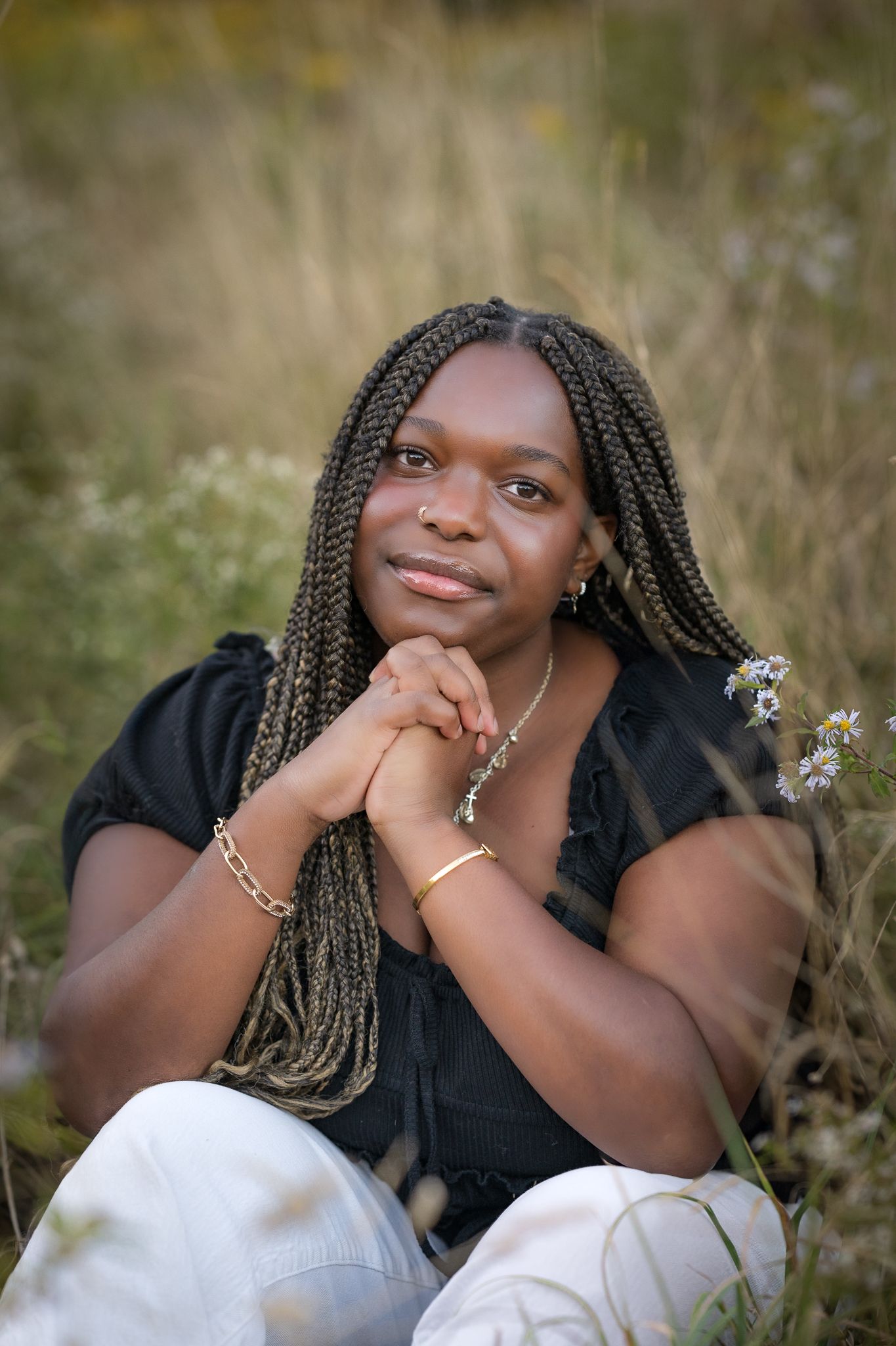 Young Black woman with braids sitting in meadow with wildflowers, hands clasped under chin.