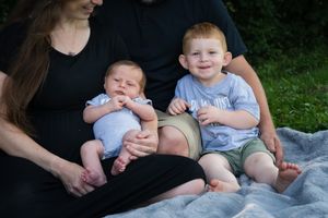 Family portrait with parents and two young boys, including a newborn, sitting on a blanket outdoors.
