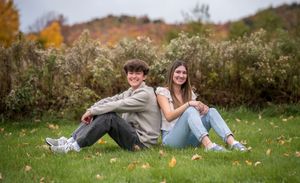 Two teens sit back-to-back on autumn grass, smiling, with colorful fall foliage in the background.