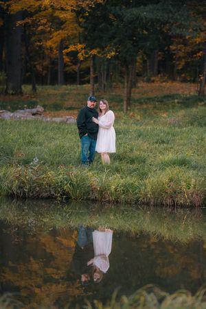 Couple embracing by a reflective pond in an autumn forest, woman in white dress and man in green jacket.