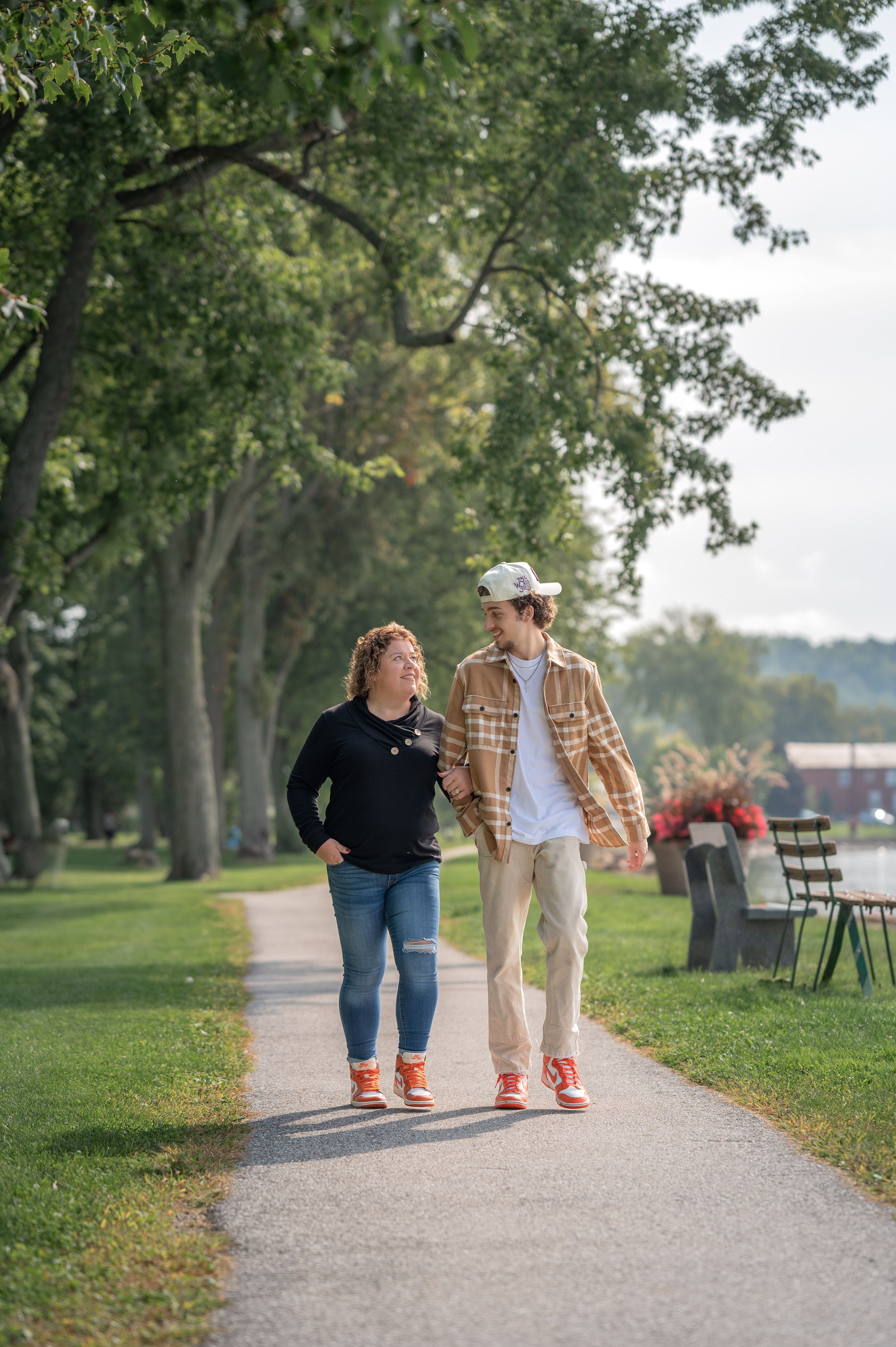 Couple walking together on a tree-lined park path, smiling at each other, wearing casual outfits and matching red sneakers.