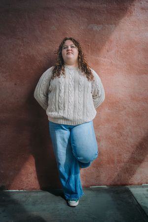 Young woman in cable knit sweater and wide-leg jeans leaning against red wall.