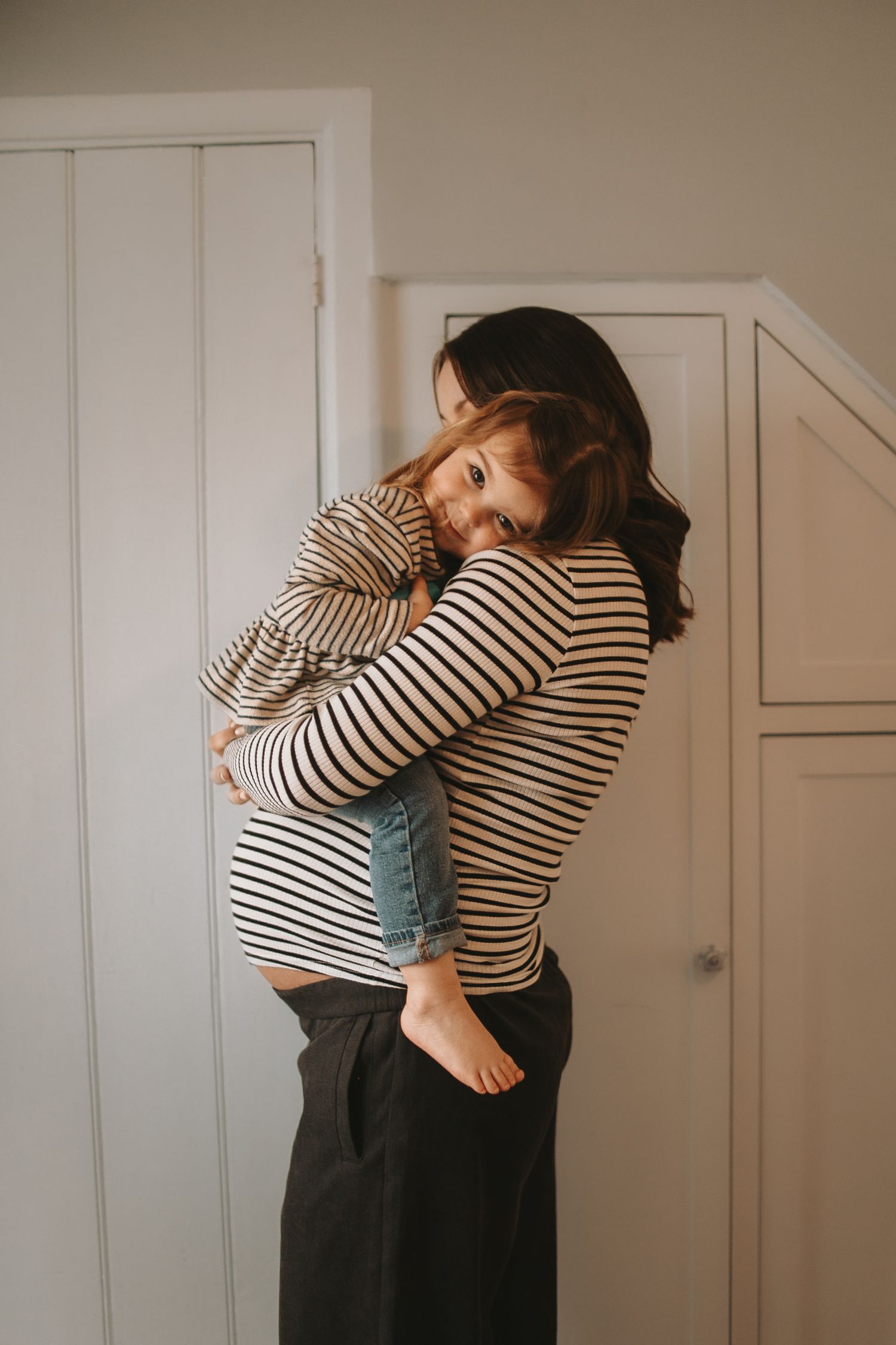 Parent in striped shirt holding small child against white cabinet door.