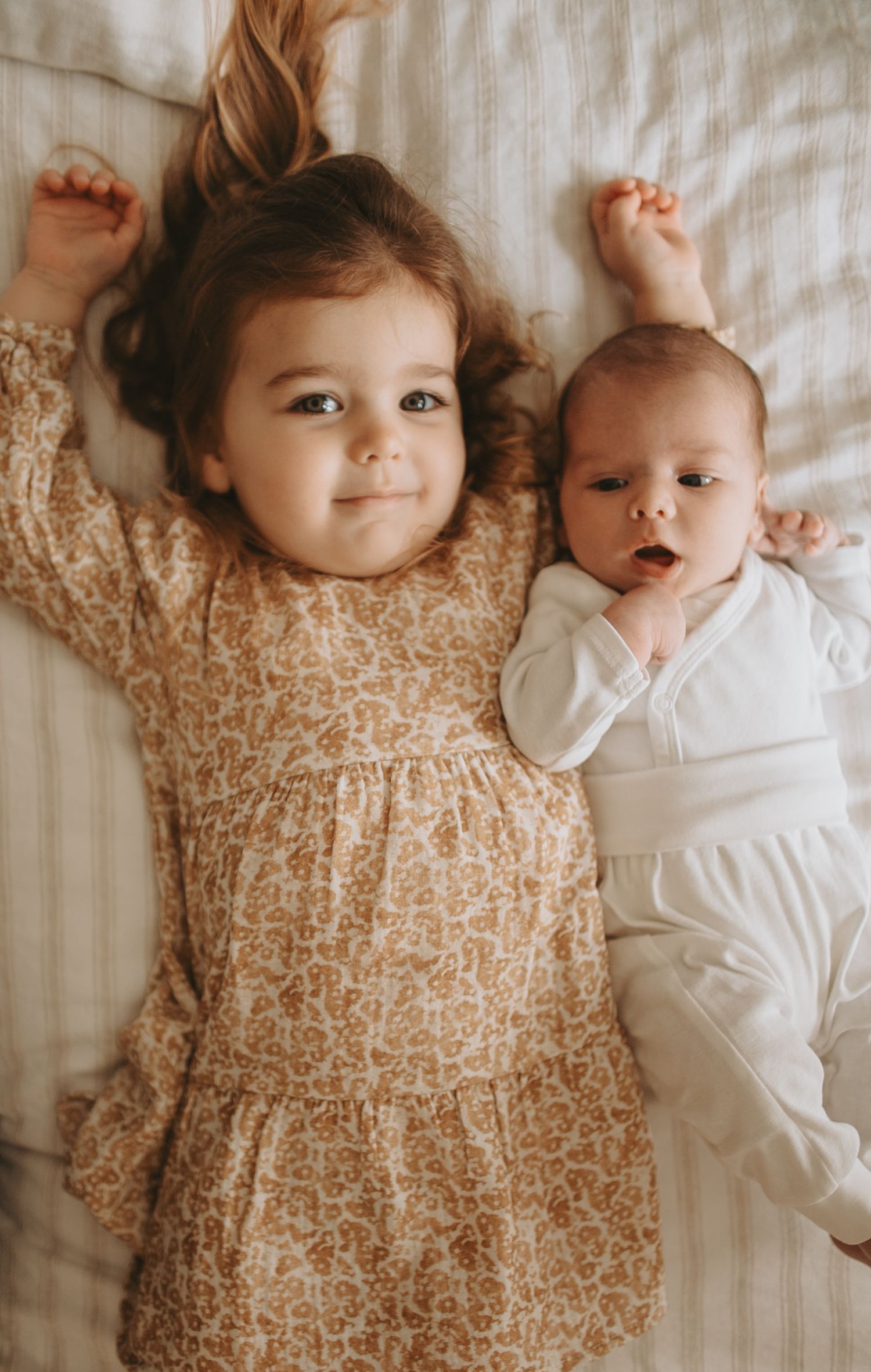 Two children lying on white bedding, one wearing a floral dress and one in white clothing.
