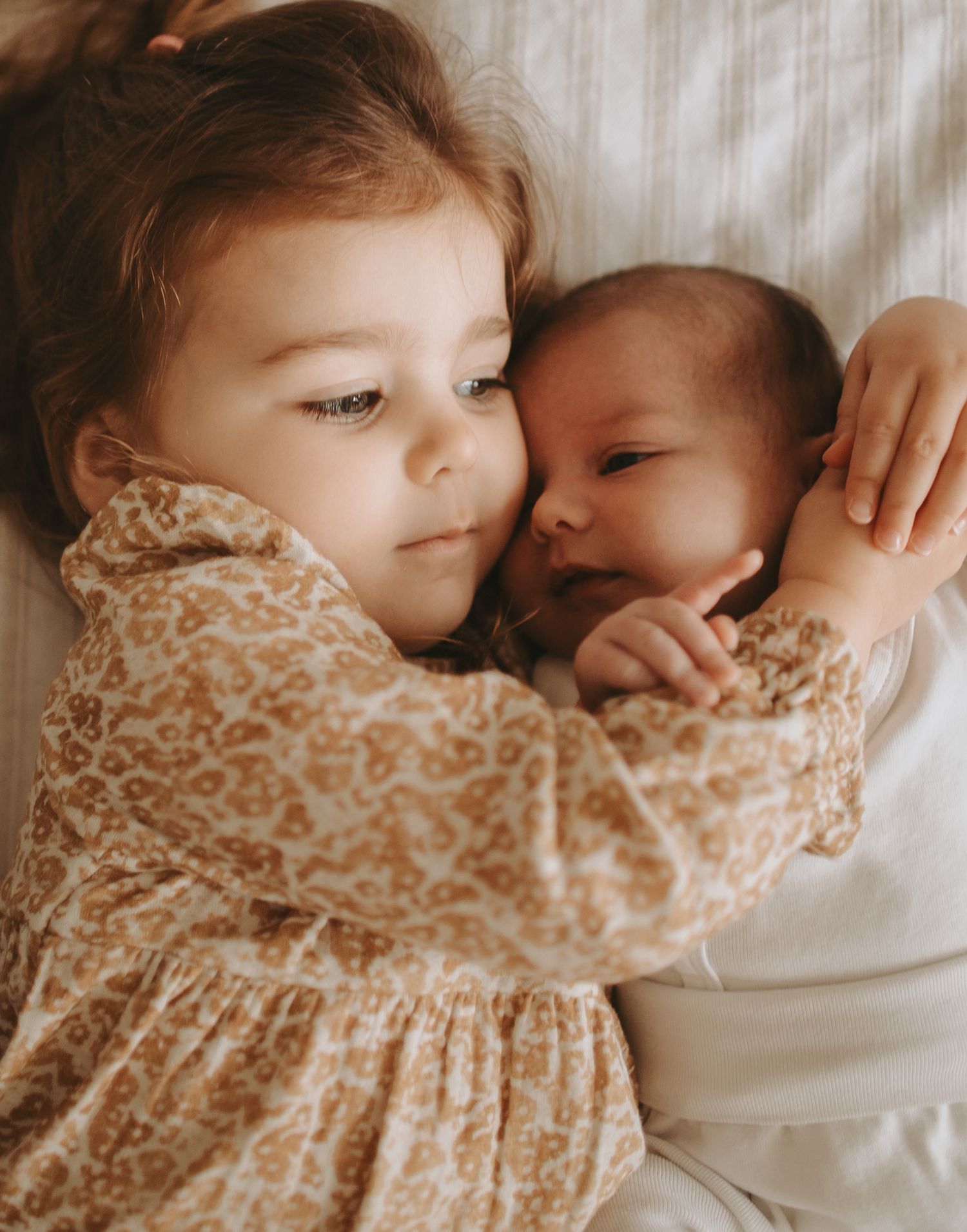Two siblings sharing a tender moment while lying on white bedding.