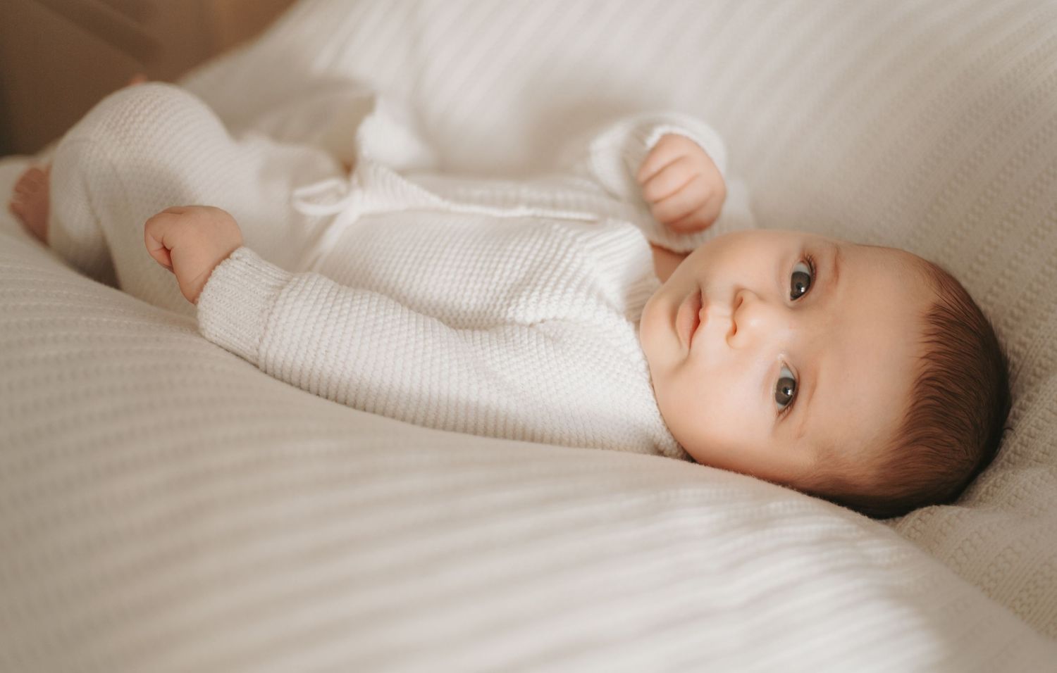 Baby in white knit clothing lying on soft white bedding during lifestyle photo session.