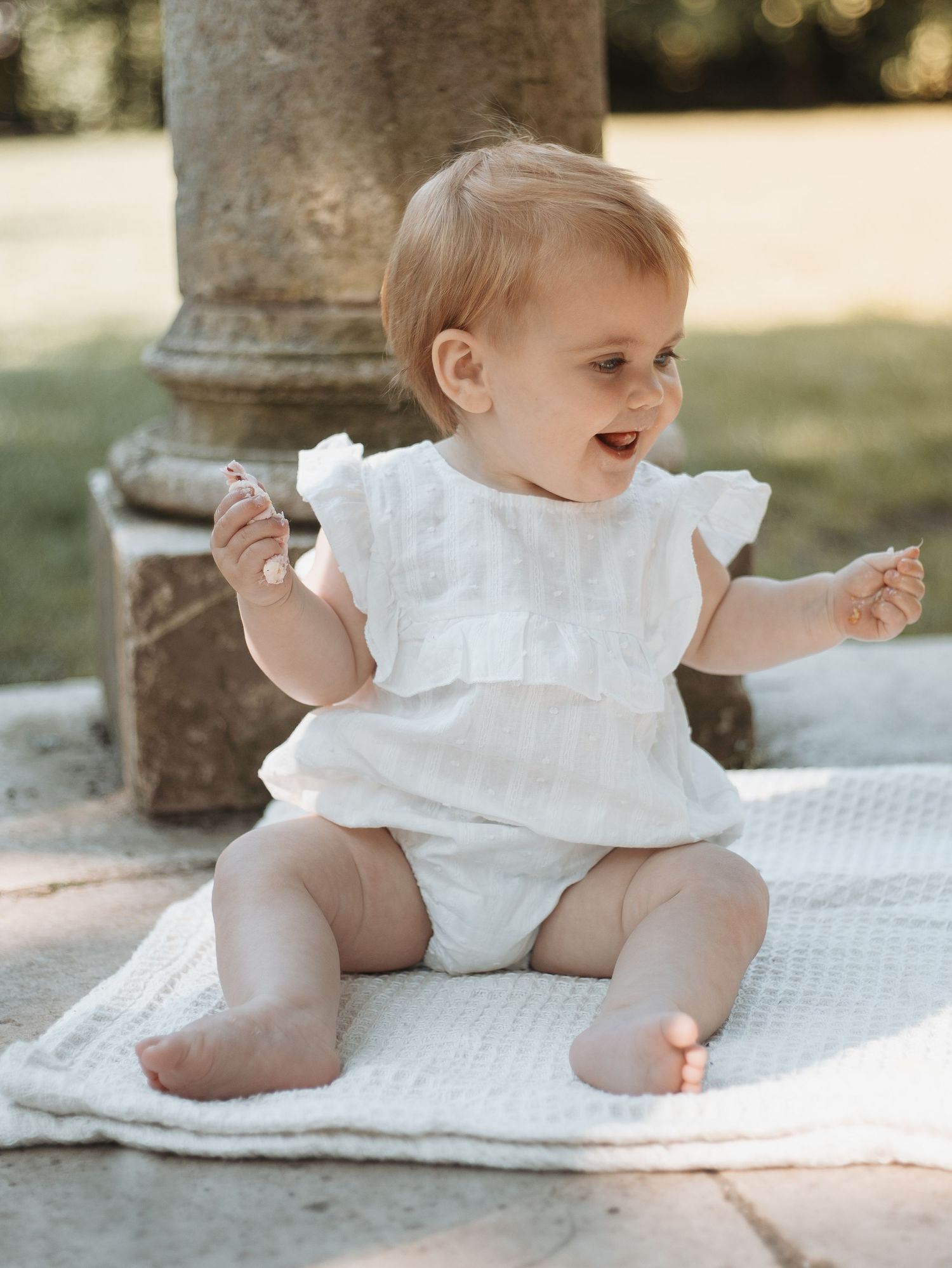 Baby in white outfit sitting on blanket outdoors near stone pillars.