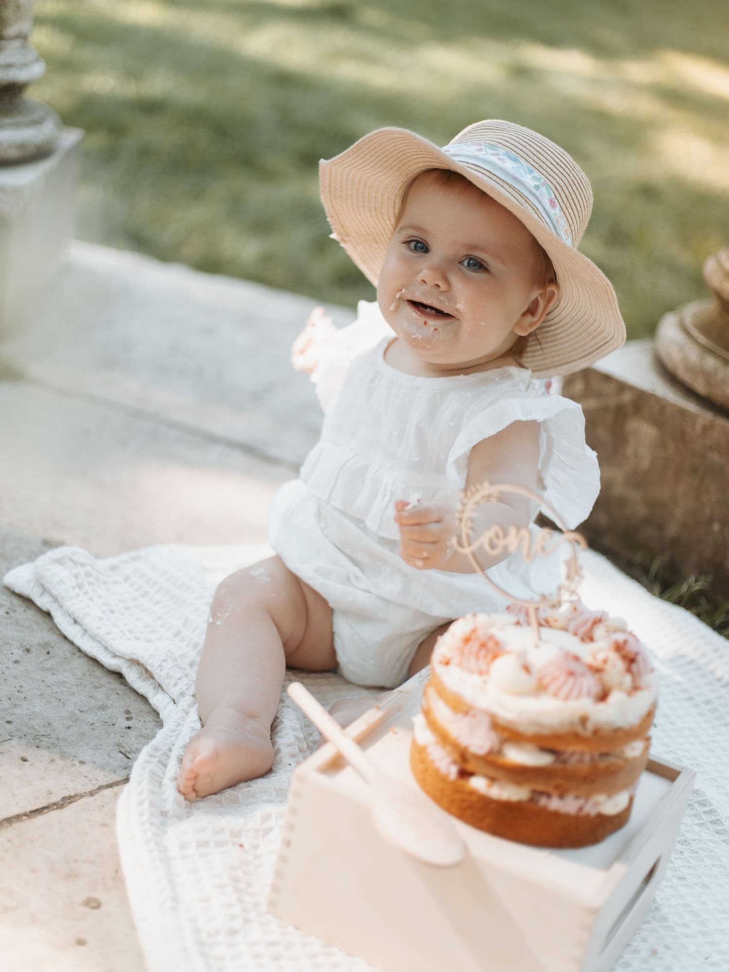 Baby in sun hat and white dress enjoying a cake smash photo session outdoors.