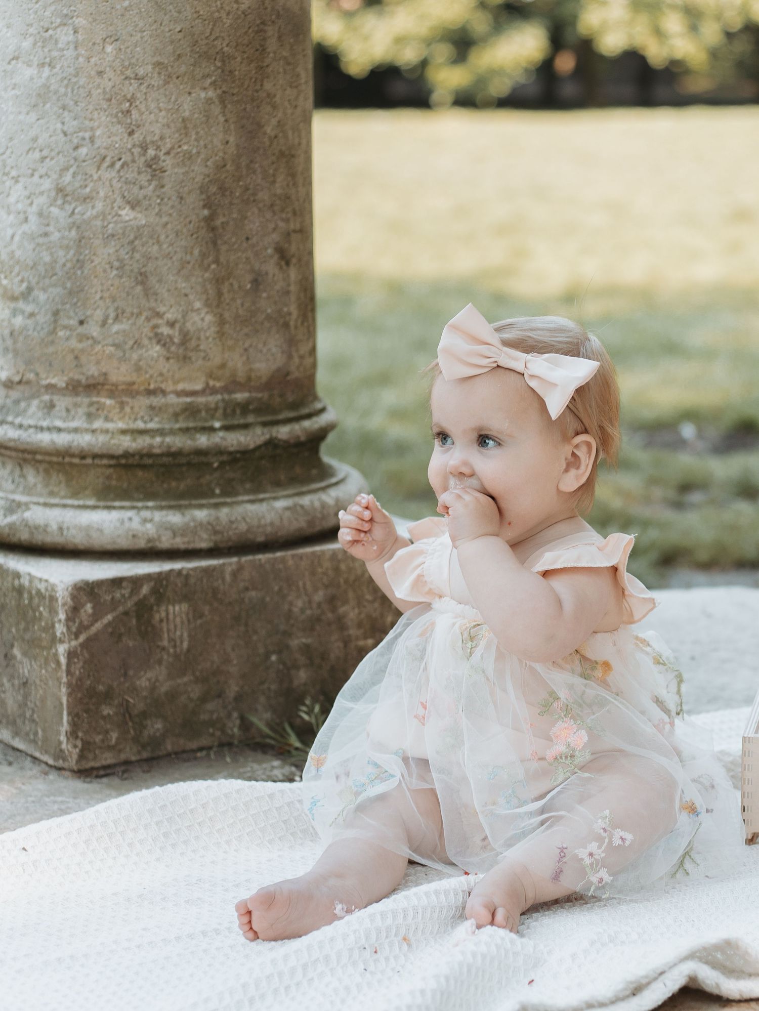Baby in frilly white dress sitting on blanket near stone architecture.