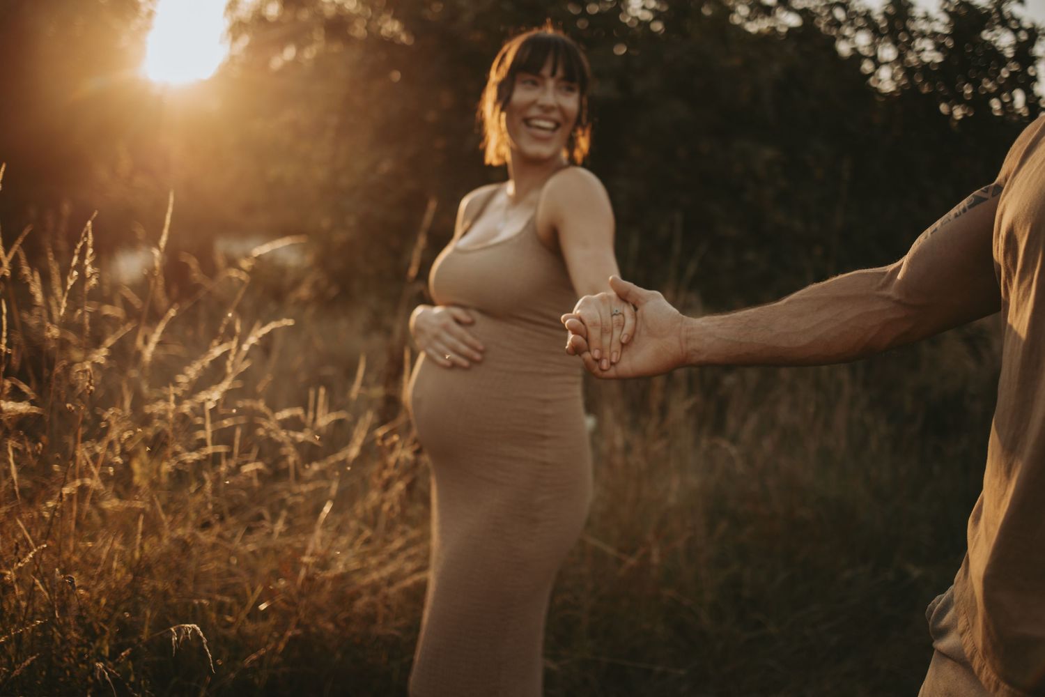 A pregnant woman in a fitted tan dress poses in golden sunlight at sunset.