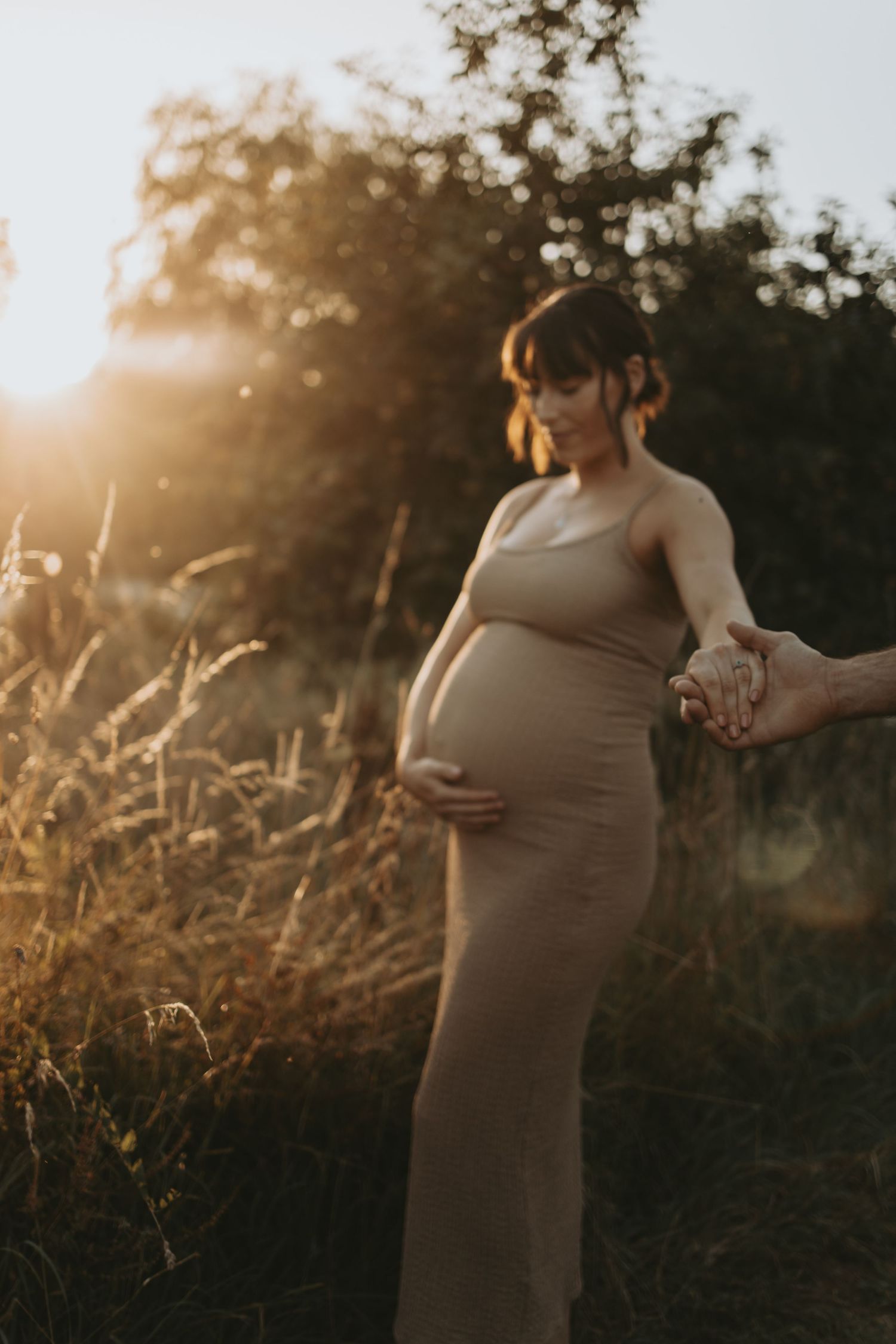 A pregnant figure in a form-fitting dress stands in golden hour light while holding someone's hand.