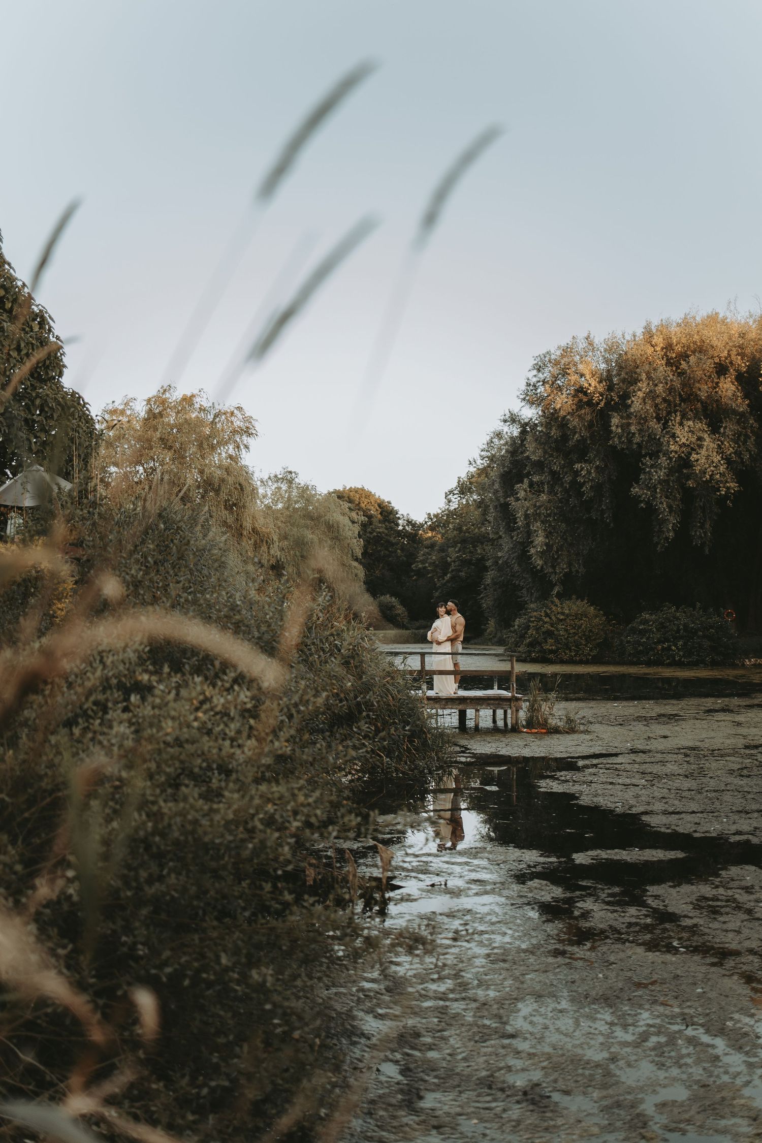 A distant couple stands on a wooden dock over a pond during sunset.