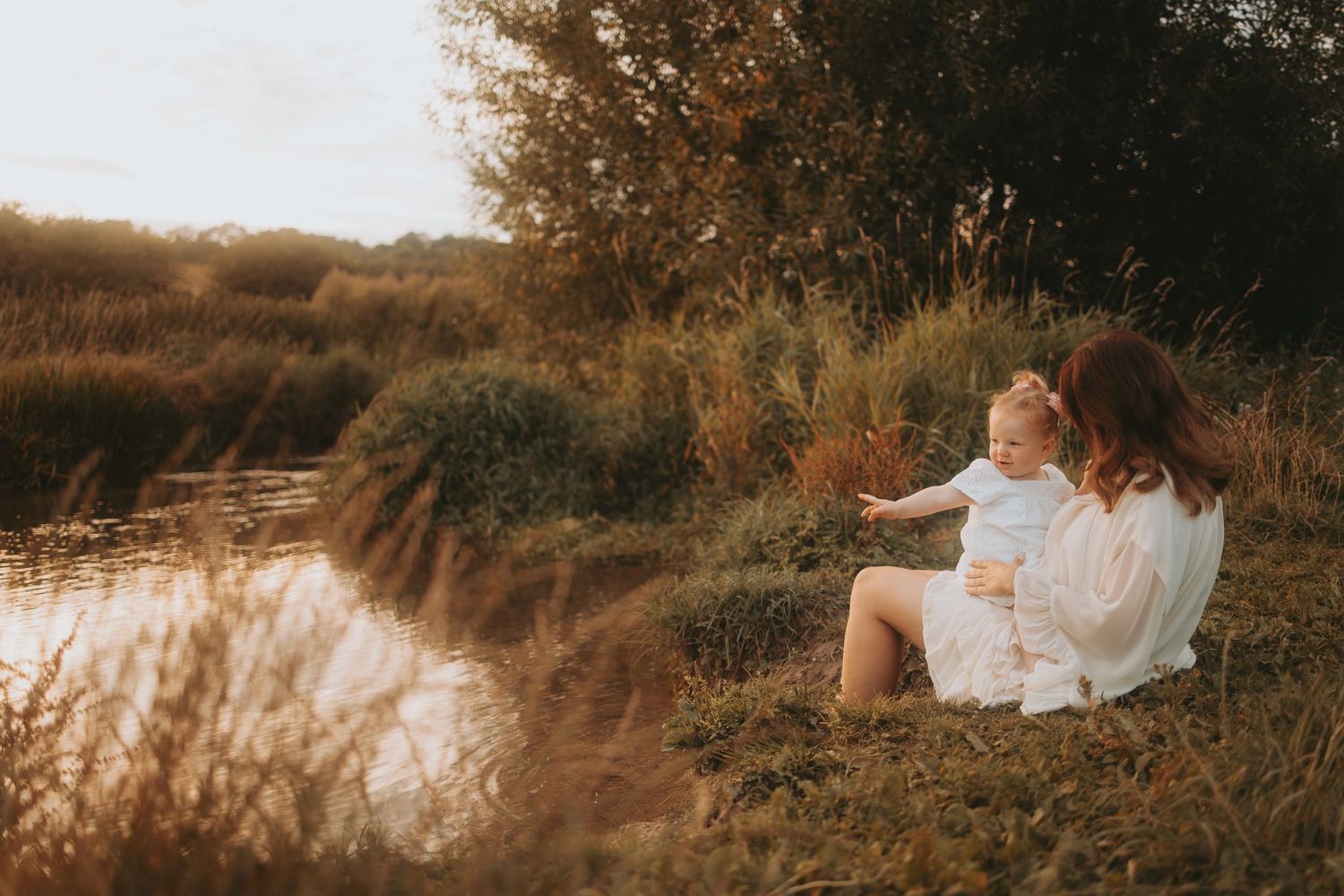 A dreamy sunset photoshoot by a lake with golden light filtering through tall grass and reeds.