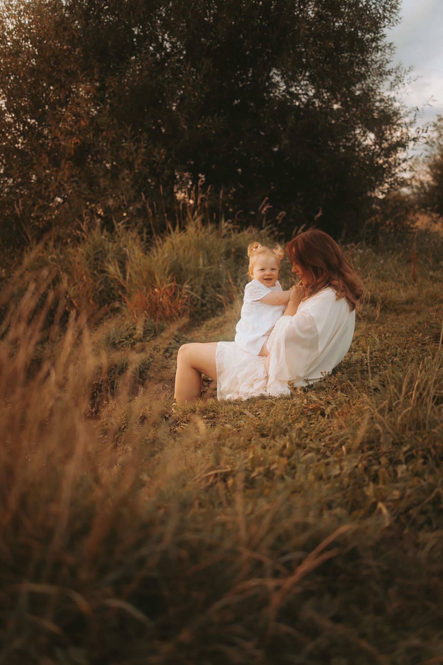 Mother and child in white dresses share a tender moment in a golden field at sunset.
