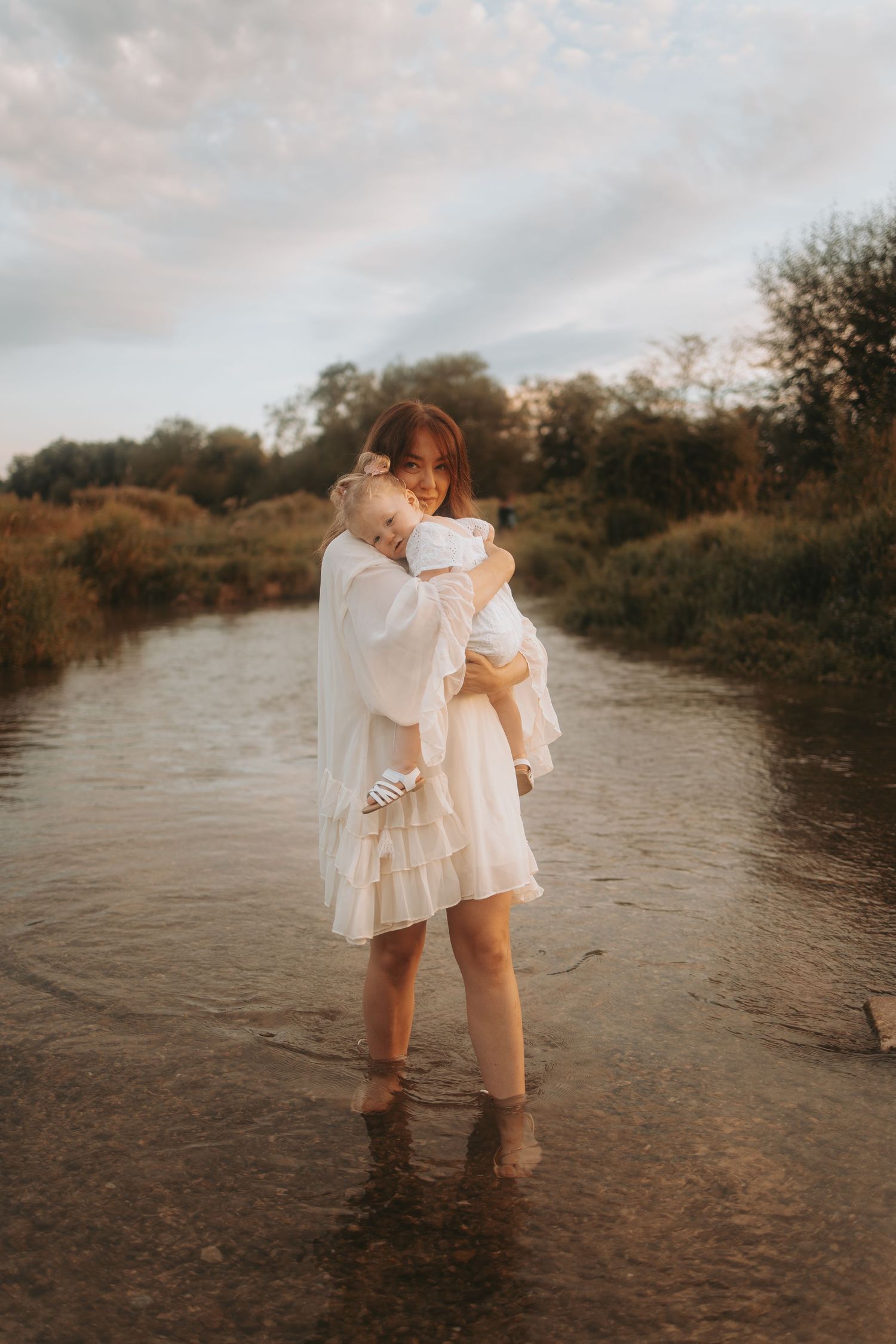 Two figures in flowing white dresses wade together in shallow river water at sunset.