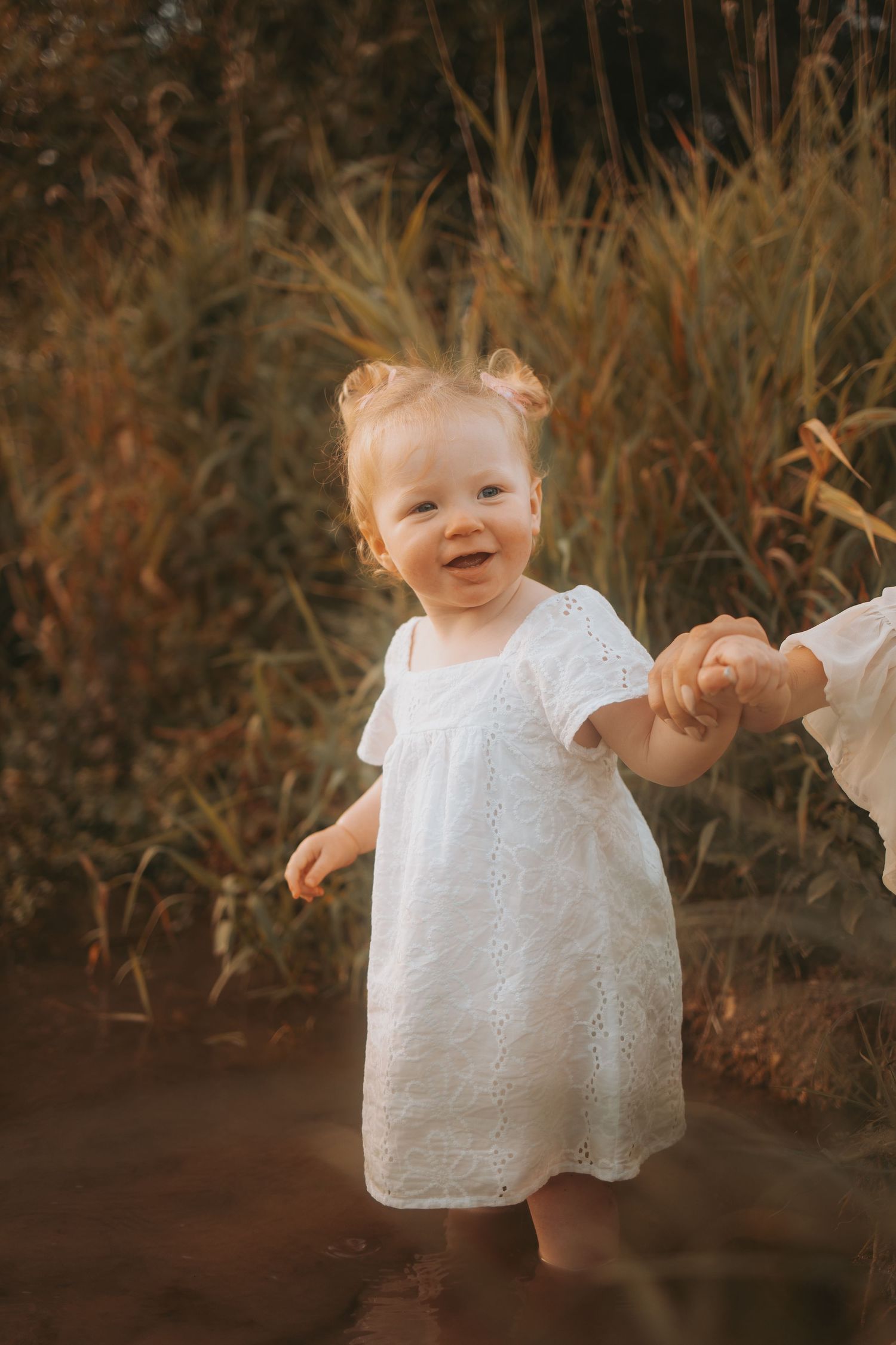 A small child in a white eyelet dress smiles and plays in a rustic outdoor setting during golden hour.
