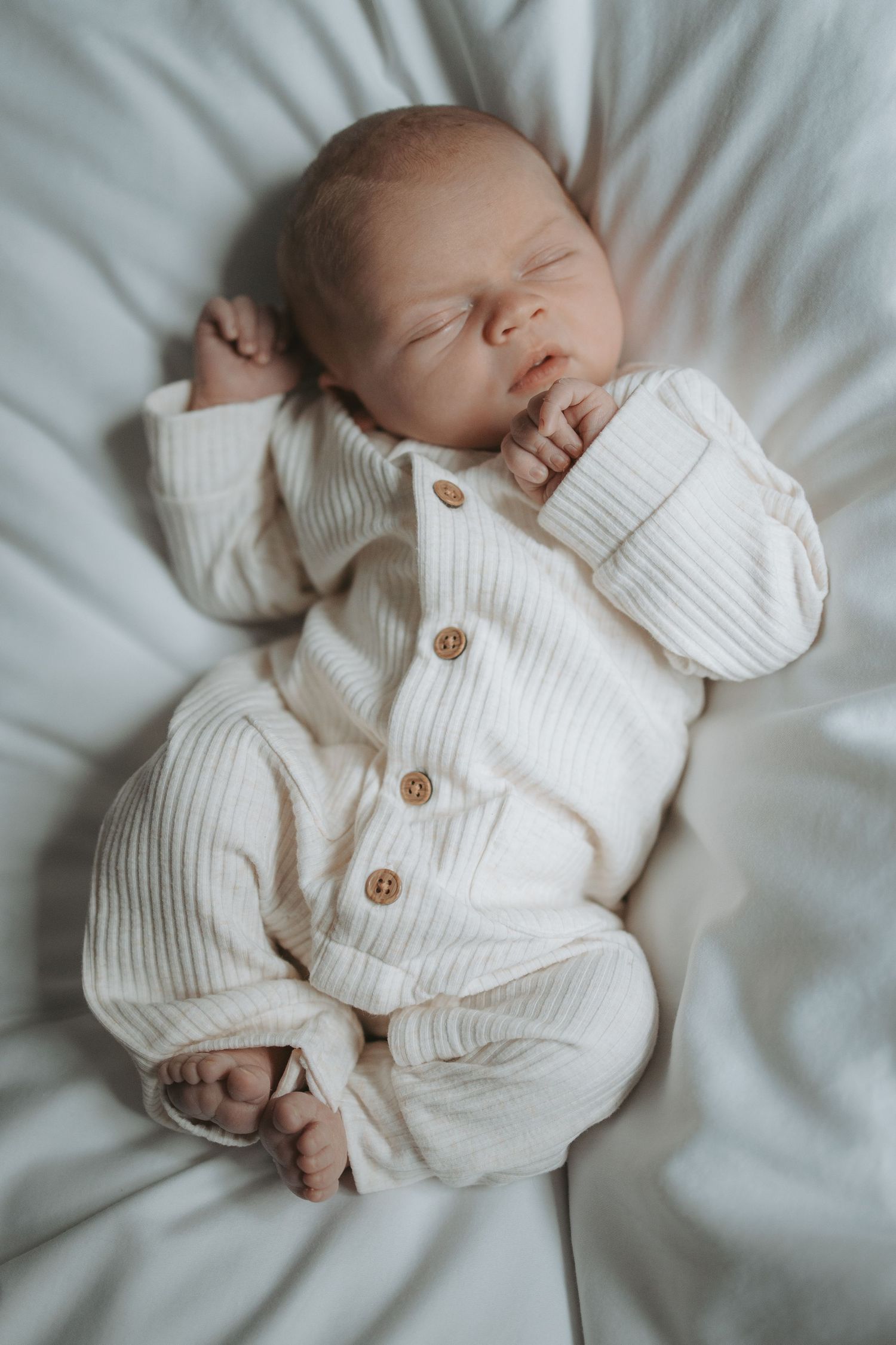 Peaceful sleeping baby wearing white knit cardigan with wooden buttons on white bedding.