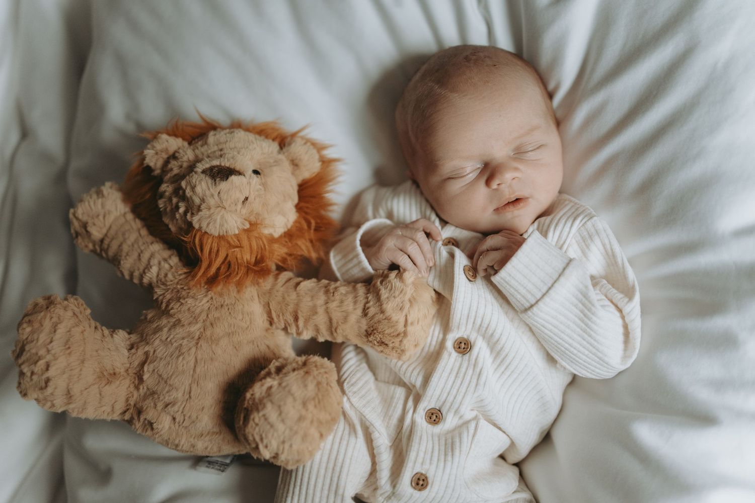 Sleeping newborn in white knit outfit lying next to plush teddy bear on white bedding.