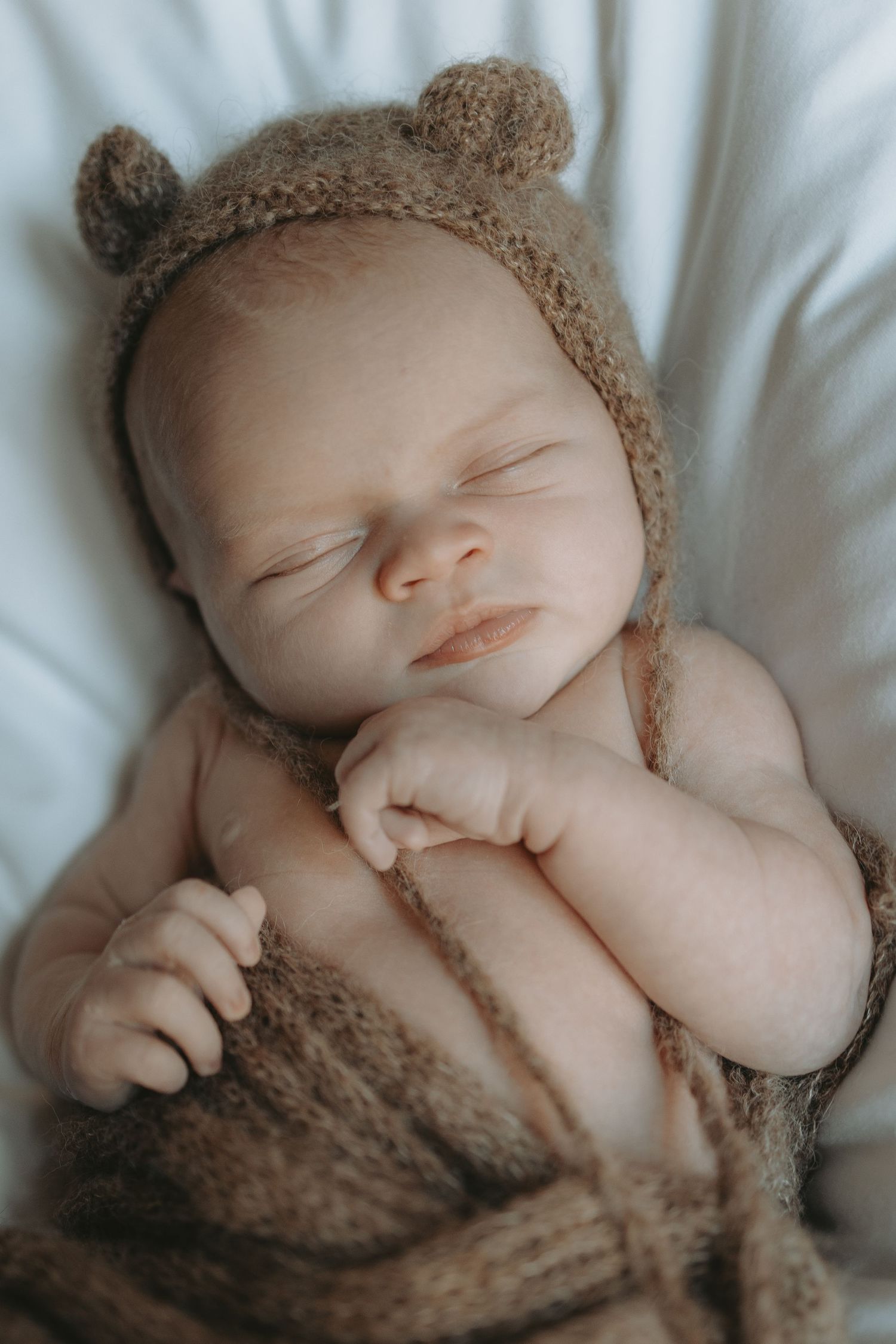 Sleeping newborn wearing knit bear bonnet wrapped in brown knitted fabric.