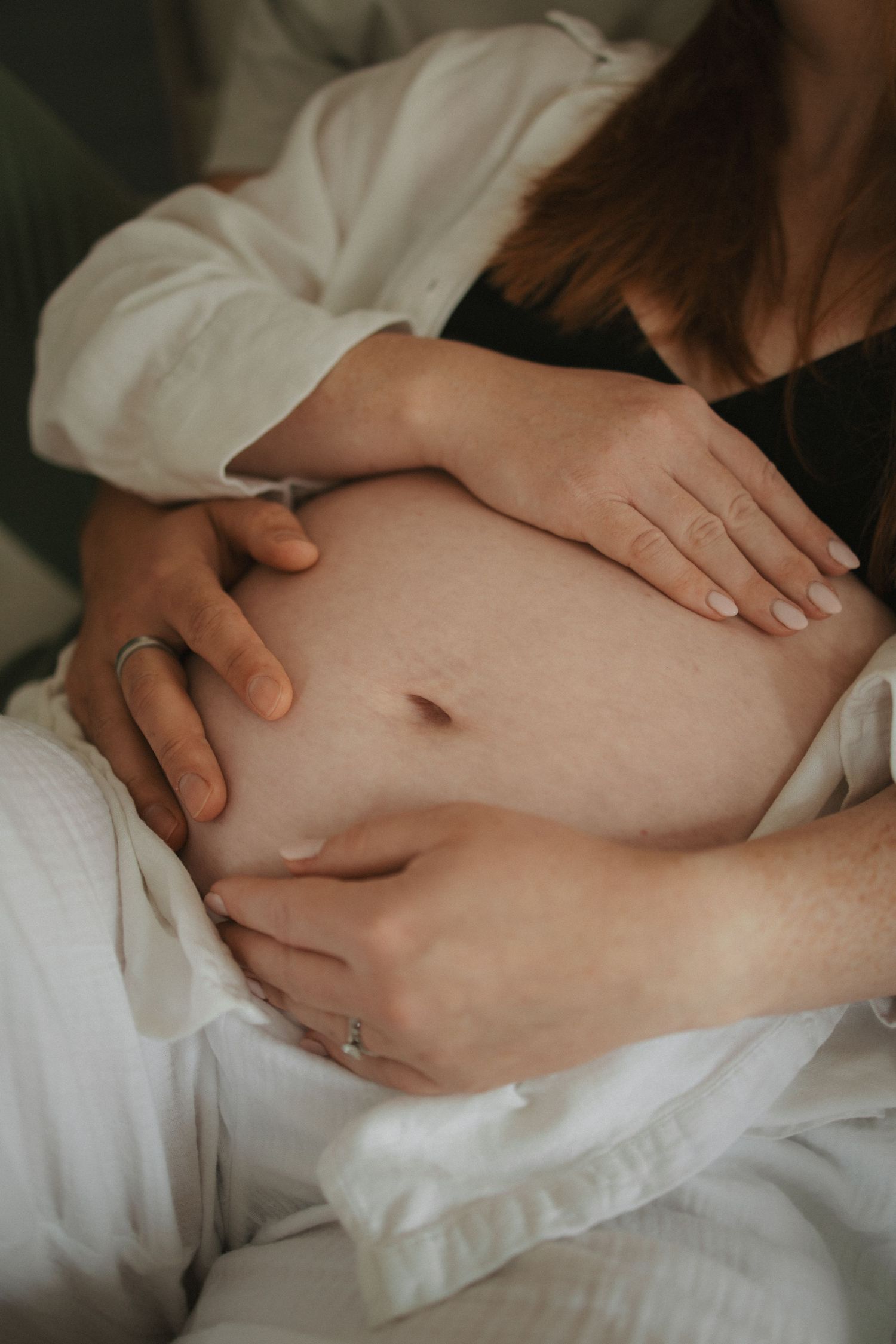 Close-up artistic shots of hands cradling pregnant belly against white sheets.