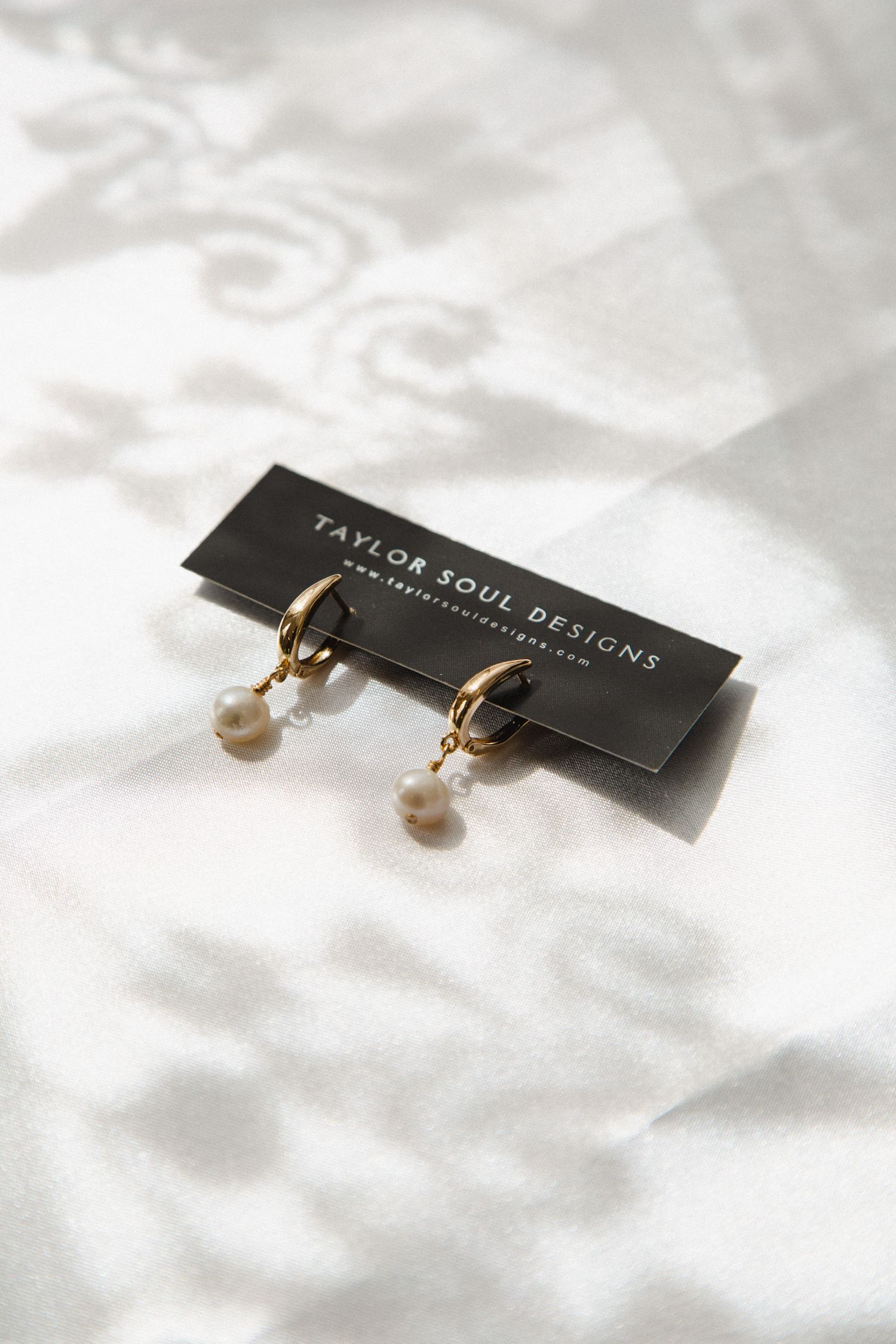 Elegant gold hoop earrings with delicate pearl drops displayed on a black Taylor Soul Designs card against white fabric.