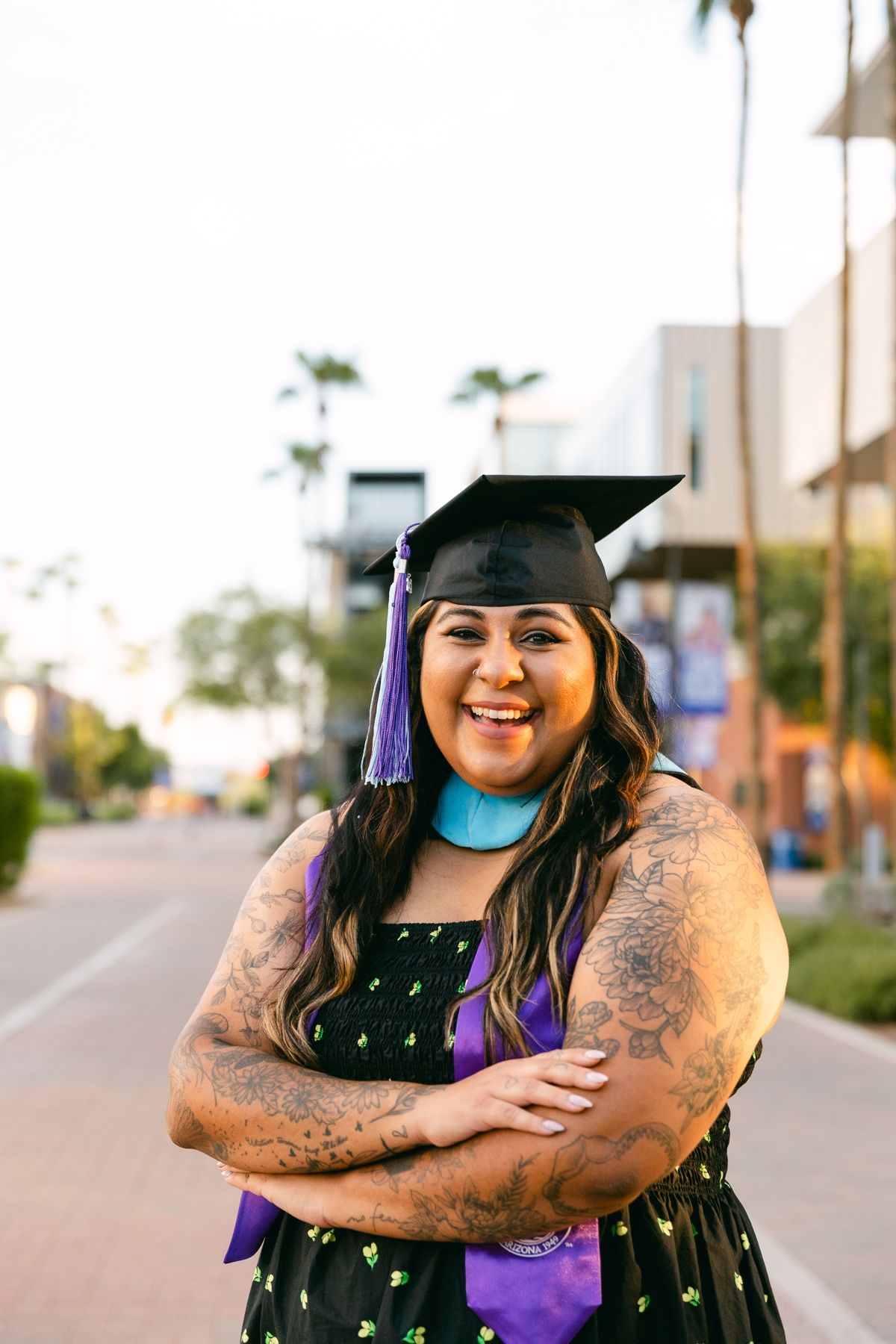 A woman wearing a black dress, a graduation cap and stole has her arms crossed and is smiling.