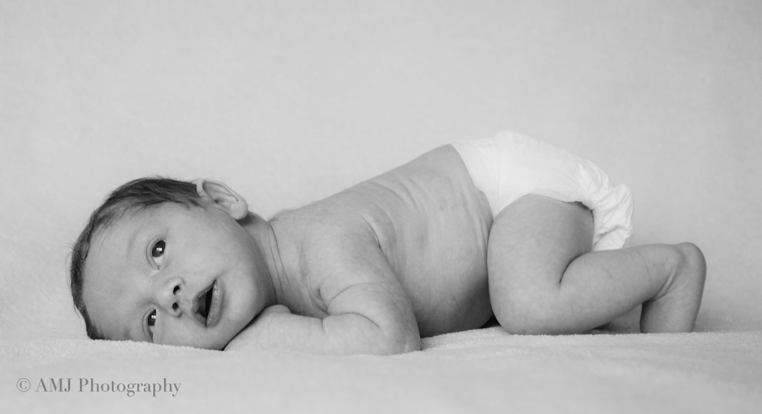 Black and white portrait of a newborn baby lying on side in a diaper on white fabric.